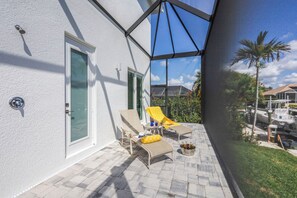 A covered outdoor patio with two lounge chairs, a small table, and a poolside area. - Huge Waterfront Home | Six Ensuites, Pool & Spa (Marco Island)