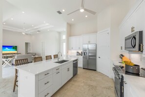 A modern kitchen with a central island, stainless steel appliances, and a dining area with a TV. - Huge Waterfront Home | Six Ensuites, Pool & Spa (Marco Island)