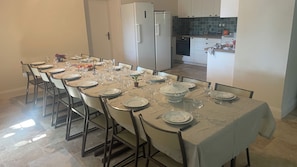 A dining area with a long table set for a meal, chairs, a refrigerator, and a kitchen area in the background.