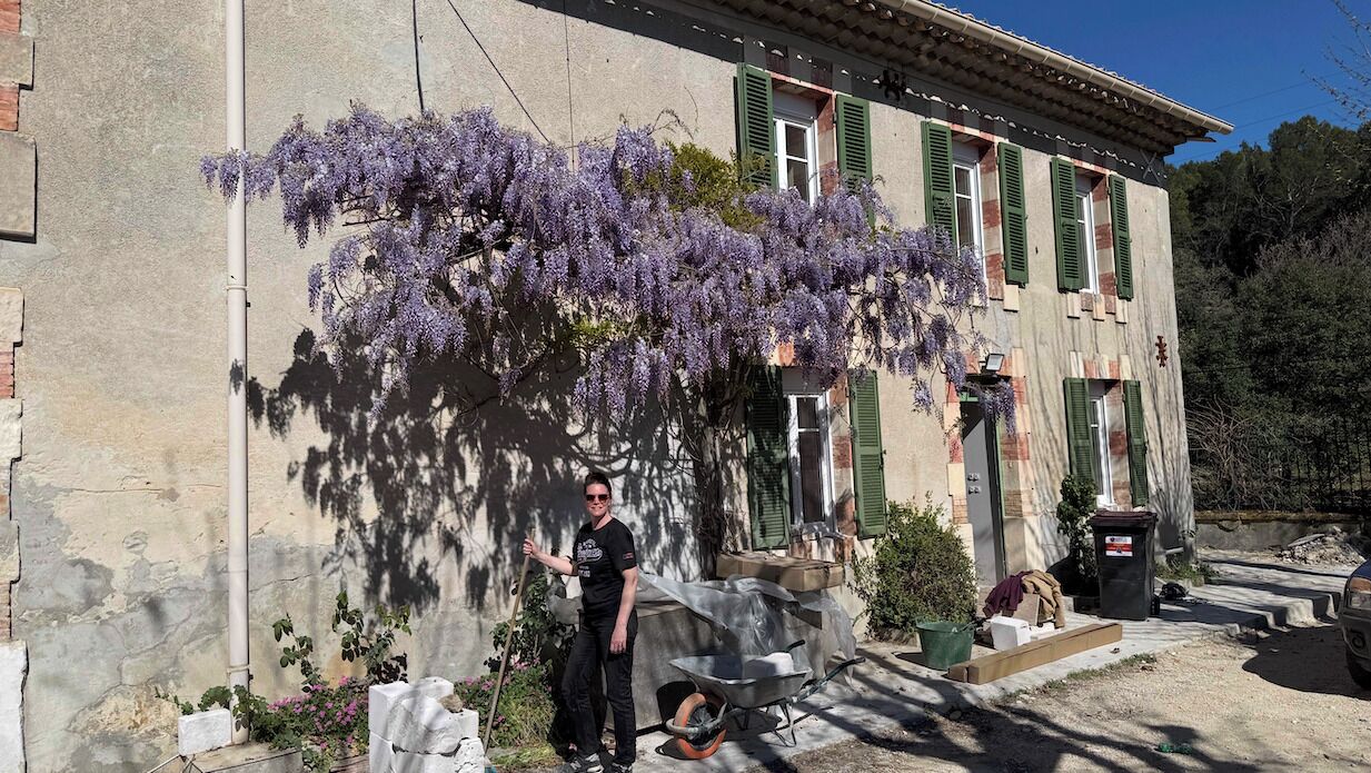 A building with a stone facade, green shutters, and a person standing next to a wheelbarrow.