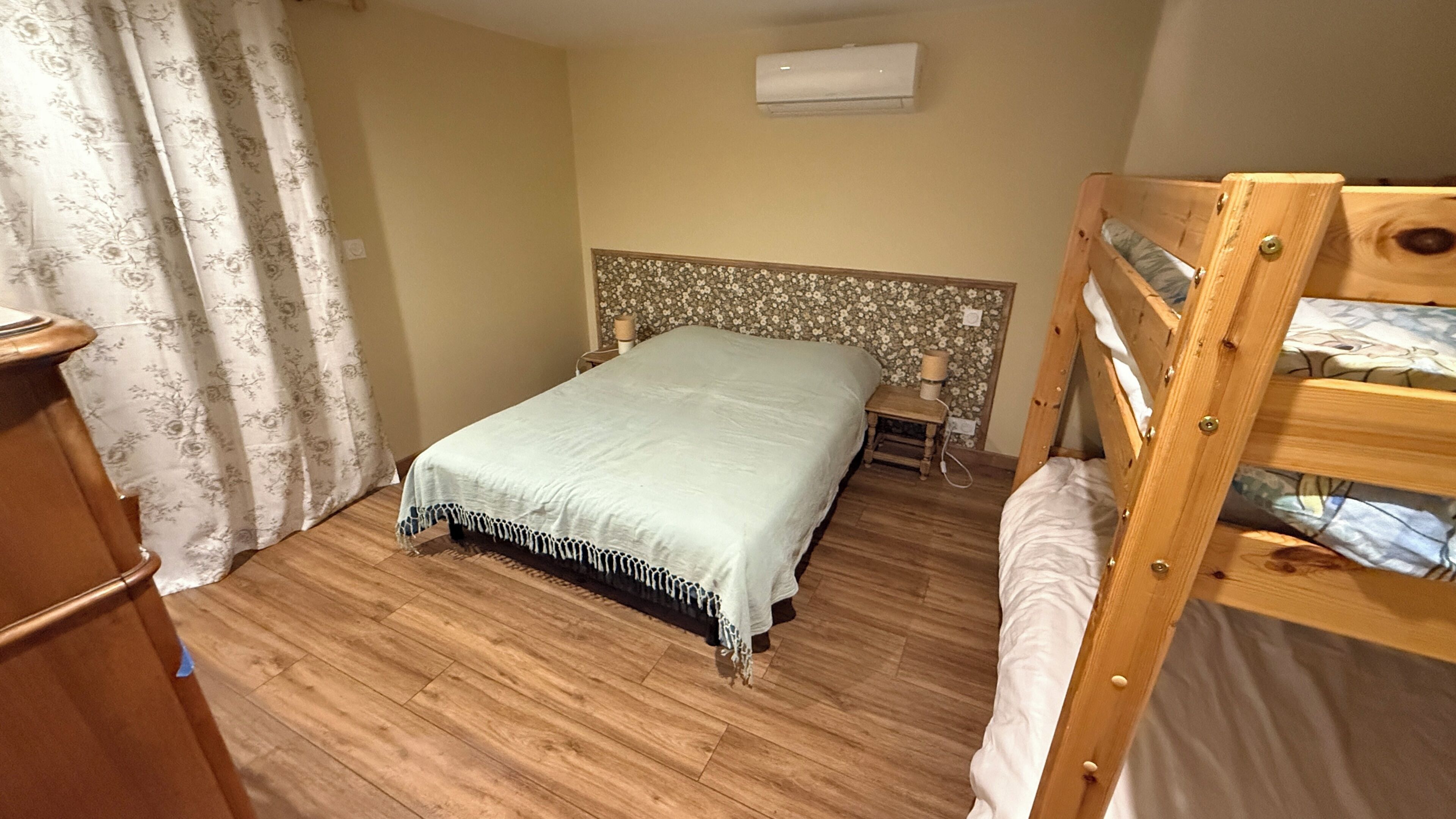 A bedroom with a wooden floor, a bed with a white bedspread, a wooden bunk bed, a nightstand, and a wall-mounted air conditioner.