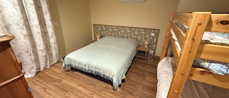 A bedroom with a wooden floor, a bed with a white bedspread, a wooden bunk bed, a nightstand, and a wall-mounted air conditioner.