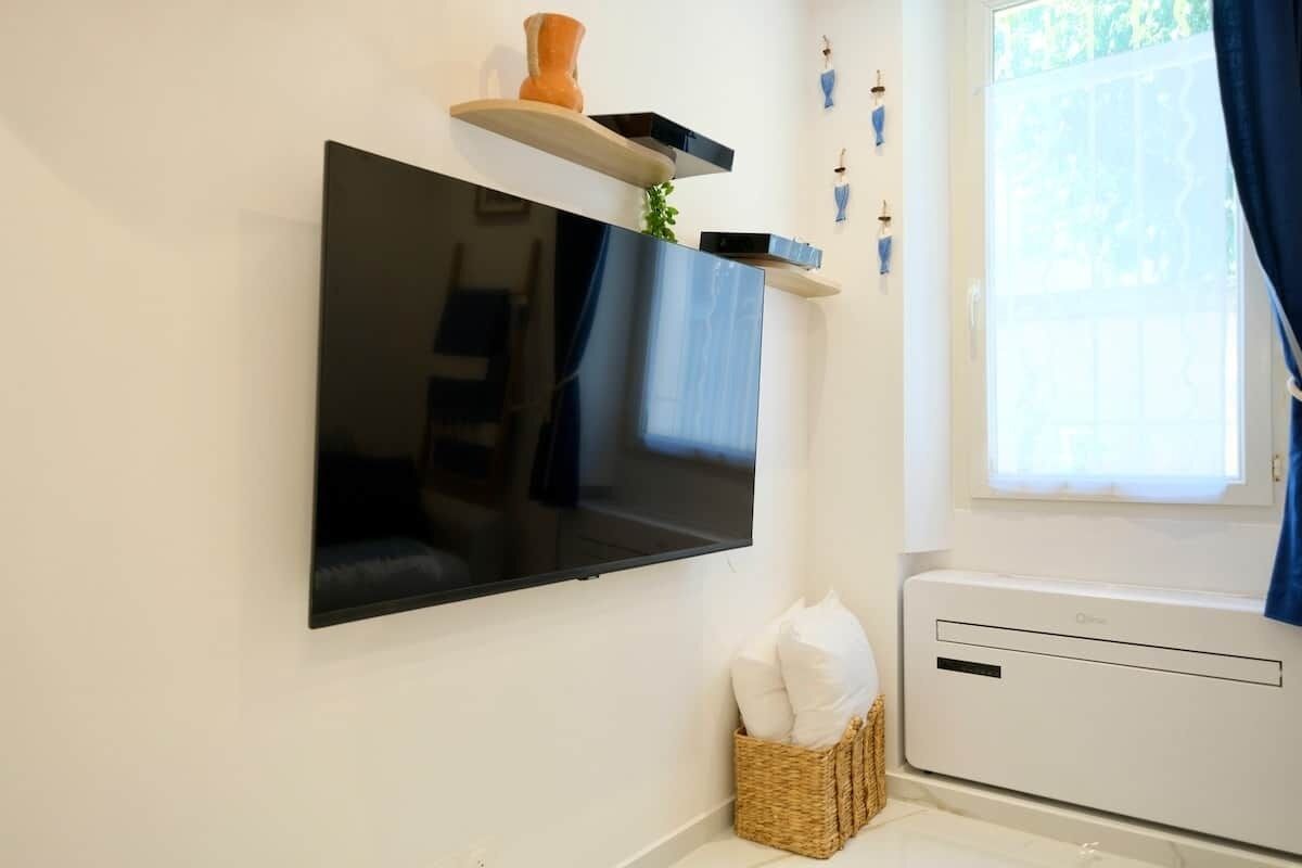 Un téléviseur à écran plat fixé au mur, un petit panier en osier contenant des coussins et une fenêtre avec des rideaux.
