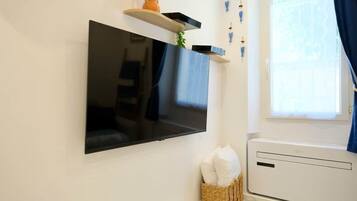 Un téléviseur à écran plat fixé au mur, un petit panier en osier contenant des coussins et une fenêtre avec des rideaux.