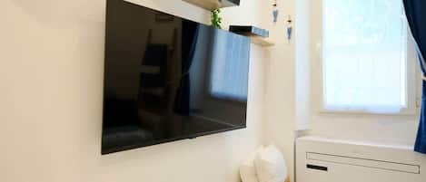 A flat-screen TV mounted on a wall, a small wicker basket with pillows, and a window with curtains.