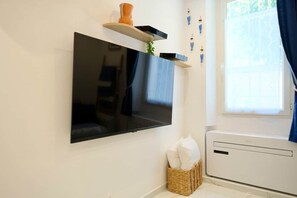Un televisor de pantalla plana montado en la pared, una pequeña cesta de mimbre con almohadas y una ventana con cortinas.