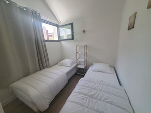 A small, clean room with two single beds, a window with curtains, a wooden shelf, and a wall-mounted light fixture. - House for rent: 41 m² - 2 bedrooms + loft - 2 terraces - direct access to the beach (Meschers-sur-Gironde)