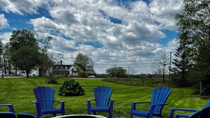 A fire pit surrounded by six blue Adirondack chairs on a gravel surface, set against a backdrop of a grassy lawn and a partly cloudy sky.