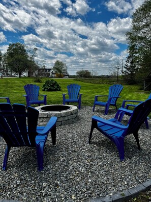 A fire pit surrounded by six blue Adirondack chairs on a gravel surface, set against a backdrop of a grassy lawn and a partly cloudy sky.