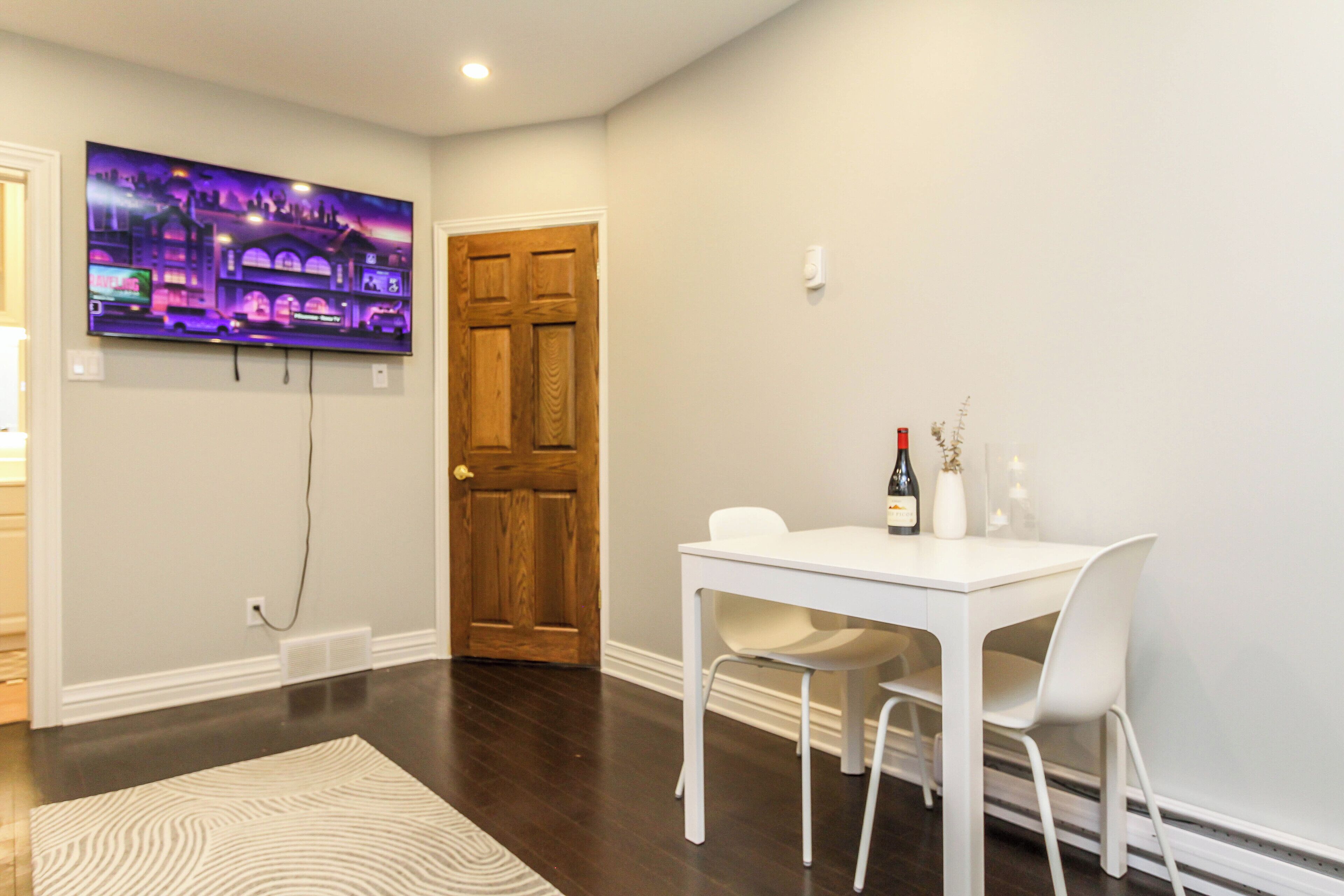 A room with a white table, two chairs, a television mounted on the wall, and a wooden door.