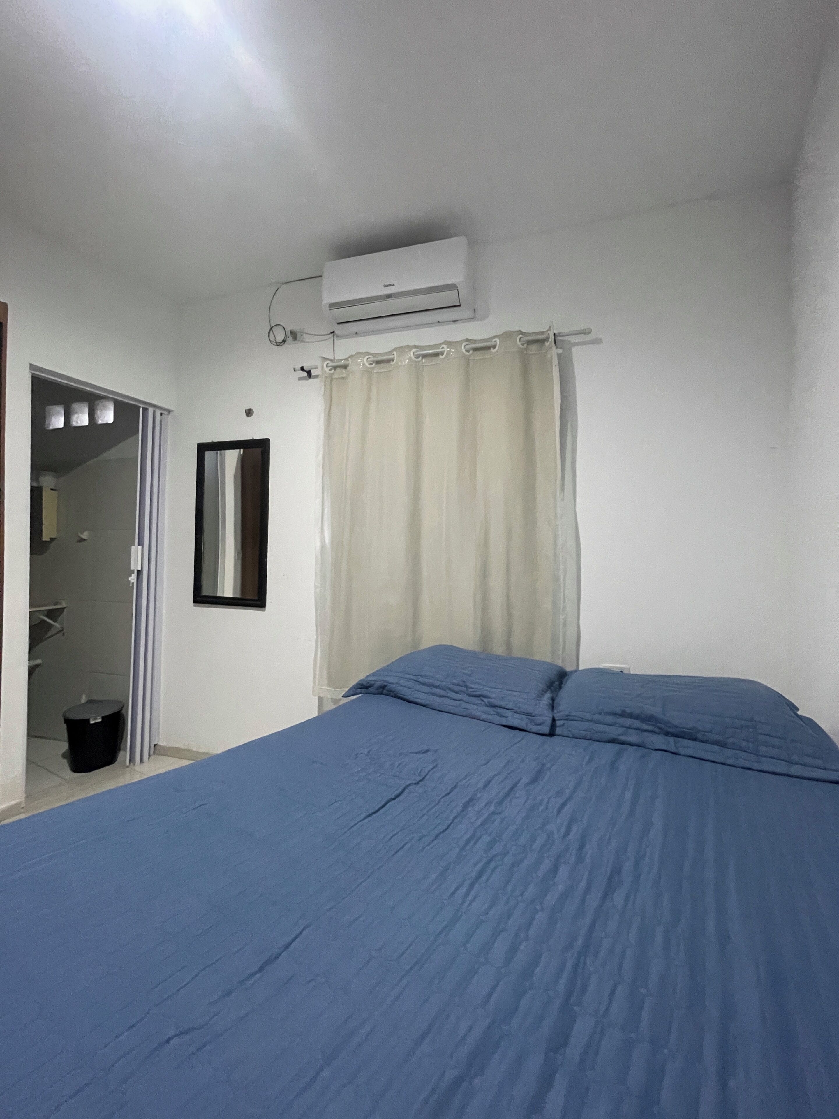 A bedroom with a bed, an air conditioning unit, a curtain, and a small trash bin.