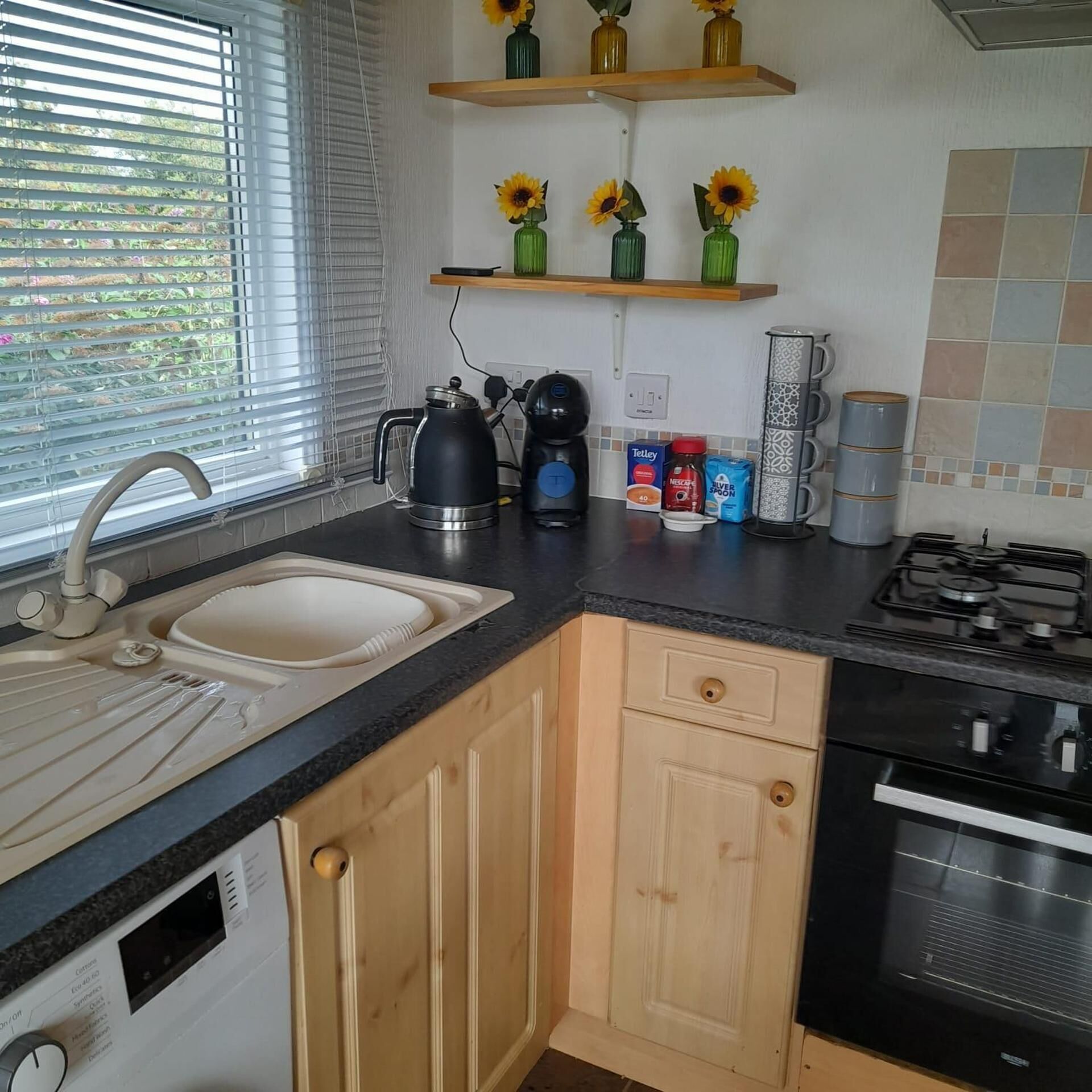 Een keuken met een spoelbak, fornuis en koffiezetapparaat. Houten keukenkastjes en een raam met zonwering.