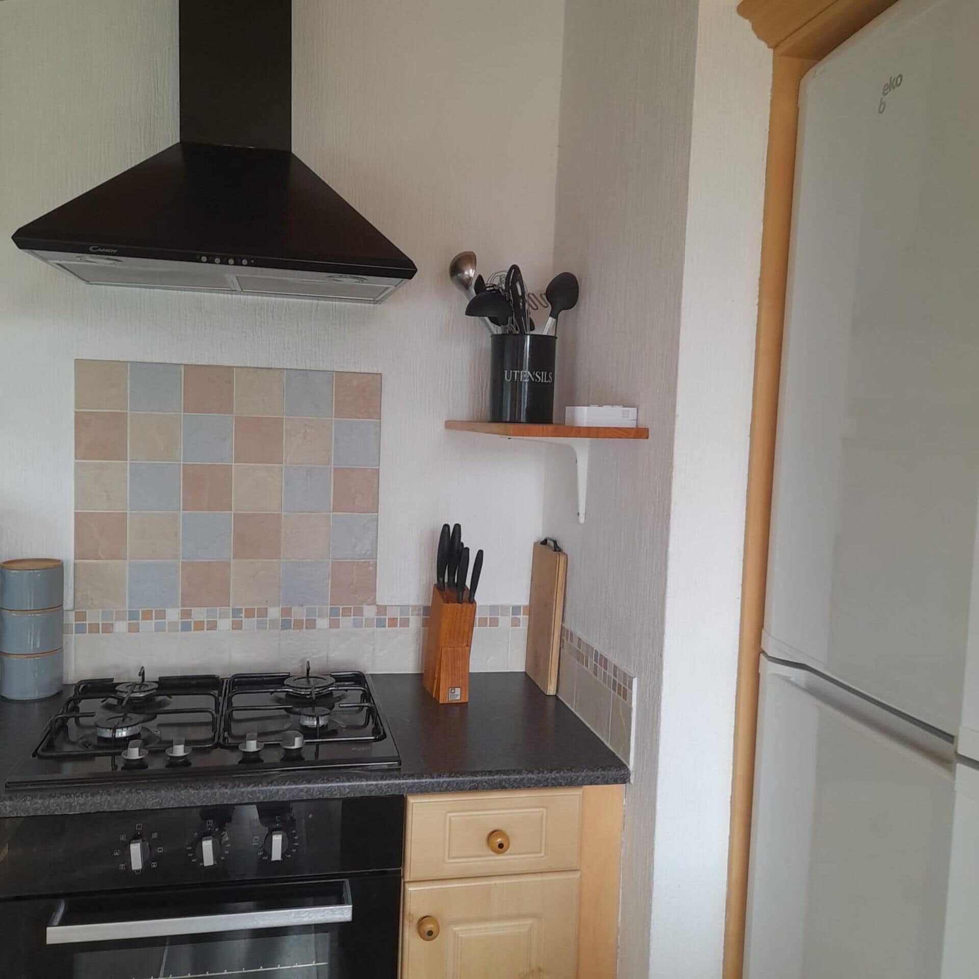 Una cocina con estufa a gas, extractor, salpicadero de azulejos, mueble de madera y refrigerador.