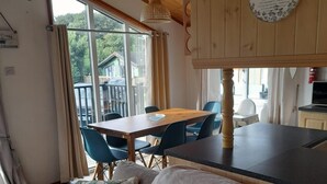 A cozy room with a wooden ceiling, a dining table with chairs, a sofa with cushions, and a view of the outside through large windows.