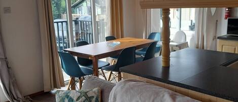A cozy room with a wooden ceiling, a dining table with chairs, a sofa with cushions, and a view of the outside through large windows.