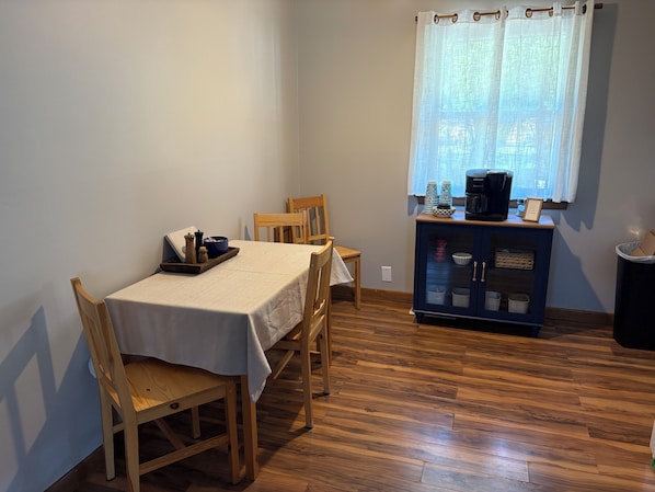 A dining area with a table set for two, wooden chairs, a cabinet with kitchen items, and a window with curtains.