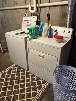 Lavadora y secadora de carga frontal con panel de control, botellas de detergente y una cesta pequeña.