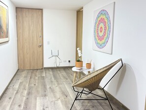 A room with a wooden door, a white wall with a colorful abstract painting, a wicker chair, a small table with a potted plant, and a white floor. - Bright 2BR in the best area of Bogotá, near Park 93 (Bogotá)