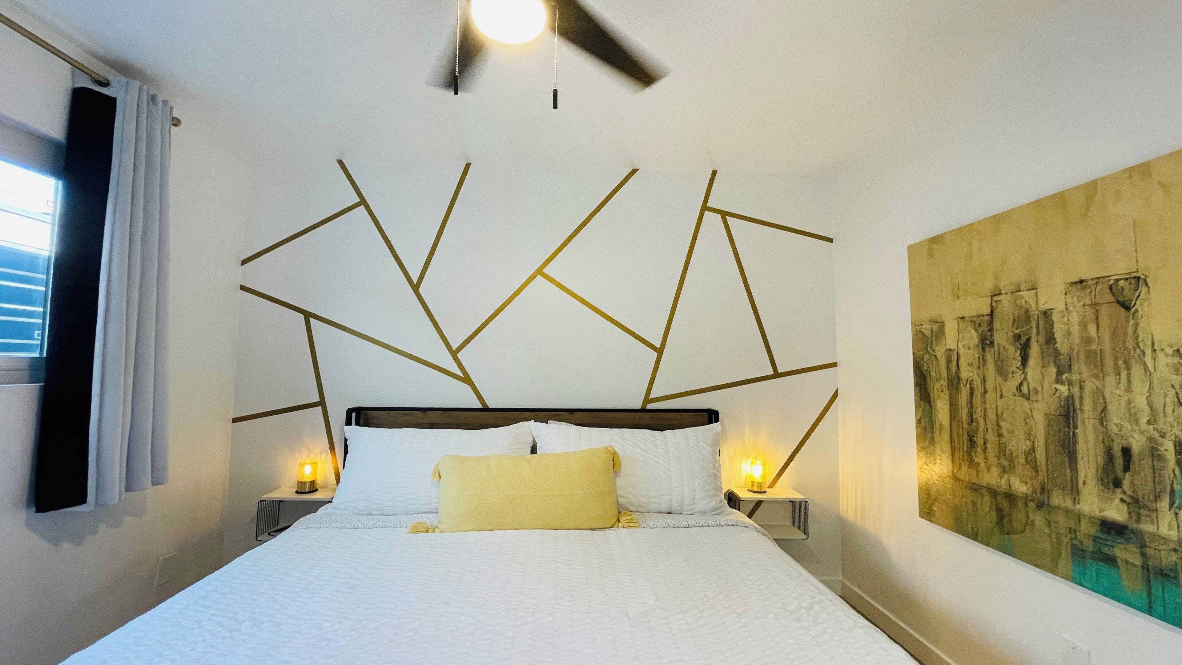 A bedroom with a modern bed, bedside lamps, a ceiling fan, and a large abstract painting on the wall.