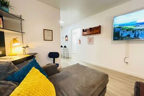A living room with a grey sofa, a yellow pillow, a desk with a lamp, and a TV on the wall. - Comfort, Convenience & Steps from Local Flavors + King Bed! (Long Beach)