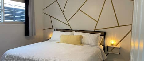 Une chambre moderne avec un motif géométrique sur le mur, un lit blanc recouvert d’un tissu texturé et des lampes de chevet.