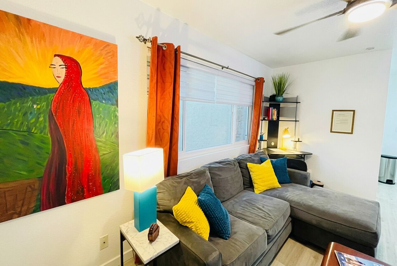 A living room with a grey sofa, a table with a lamp, a painting on the wall, and a window with curtains.