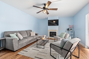 A modern living room with a grey sofa, a coffee table, a fireplace, and a ceiling fan. - New Smyrna Beachside 2BR 3 Bath Condo Balcony (New Smyrna Beach)
