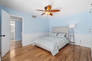 A bedroom with a bed, a ceiling fan, a bedside lamp, and a door leading to a bathroom. - New Smyrna Beachside 2BR 3 Bath Condo Balcony (New Smyrna Beach)