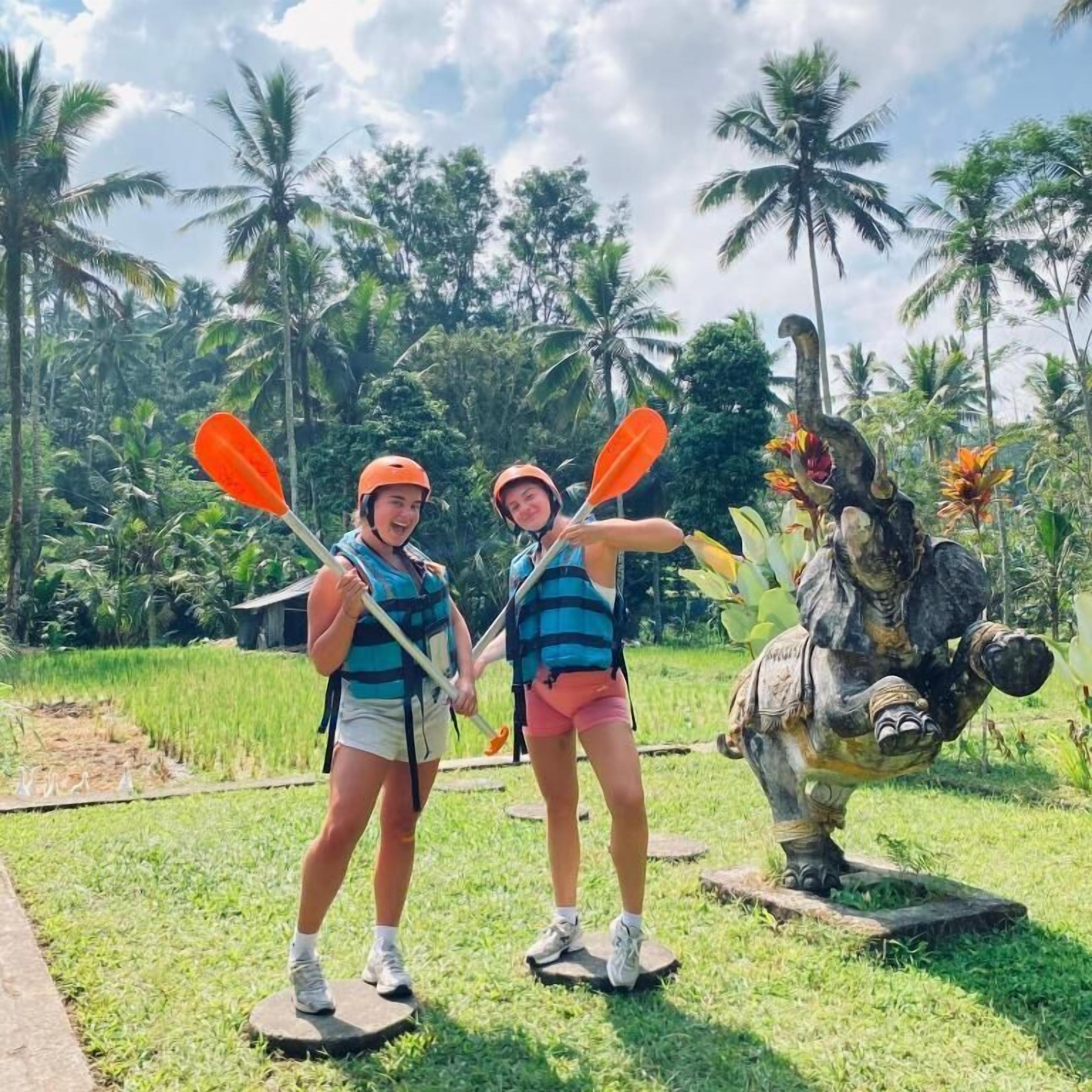 Dos personas en una plataforma sosteniendo remos, una escultura de un caballo y un fondo tropical con palmeras.