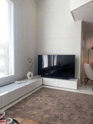 A modern living room with a flat-screen TV mounted on a white cabinet, a large window with blinds, a beige carpet, and a grey armchair. - Scandinavian Arctic Retreat in Rovaniemi – 3BR & Sauna (Rovaniemi)