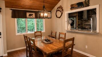 A dining area with a wooden table and chairs, a chandelier, a window with a view of trees, and a kitchen visible through the window.