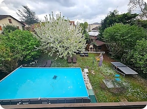 Una piscina con una terraza alrededor, un área de patio con mesa y sillas, y un jardín con árboles en flor.