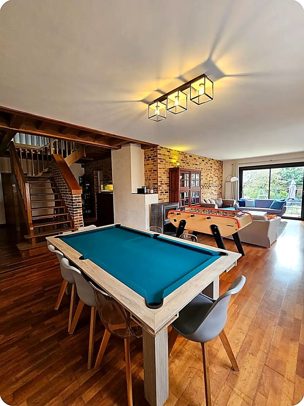 Uma mesa de bilhar em um espaço com parede de tijolos, uma mesa de futebol de mesa e uma escada.