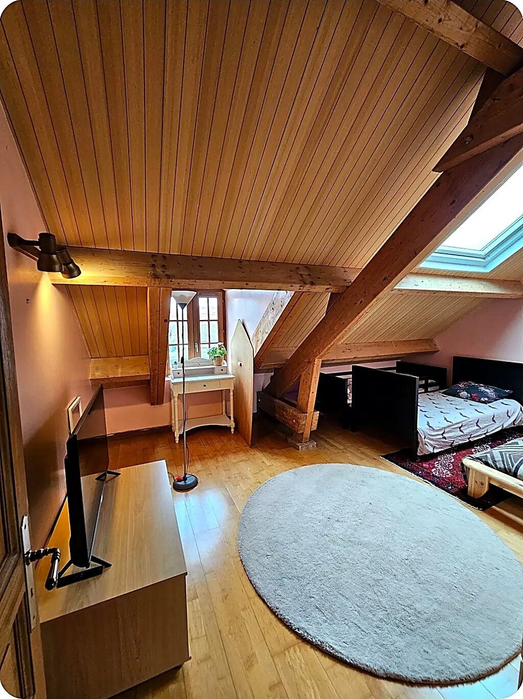 Um quarto de sótão com claraboia, vigas de madeira, uma escrivaninha, uma cama e um tapete redondo.