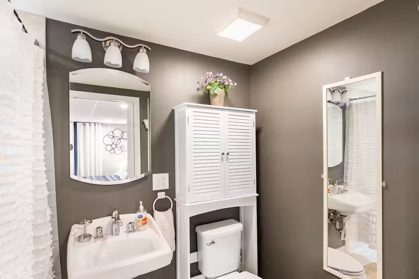 A bathroom with a white sink, toilet, and vanity, a mirror, and a cabinet with a vase of flowers.