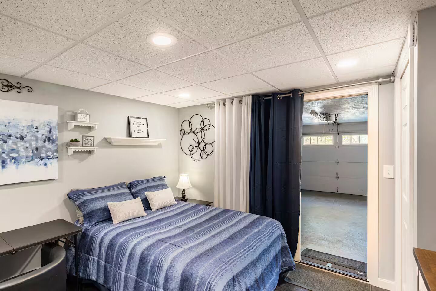 Une chambre à coucher comprenant un lit, des tables de chevet, une étagère murale décorative, un luminaire au plafond et une vue sur un garage.
