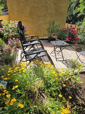Eine Holzterrasse mit Gartenmöbeln, umgeben von Kübelpflanzen und blühenden Sträuchern.