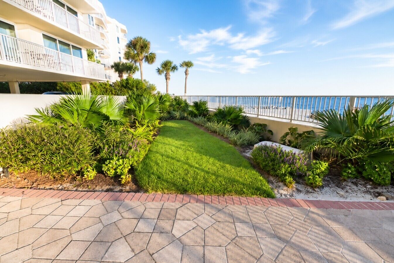 A landscaped garden with a well-maintained lawn, various shrubs, and palm trees, leading to a balcony overlooking a body of water.