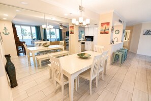 A spacious dining area with a large table and chairs, a kitchen in the background, and a view of the outdoors. - Waterview Towers 104 Harbor Views & Resort Living & Boat Slip Option Sleeps 8 (Destin)