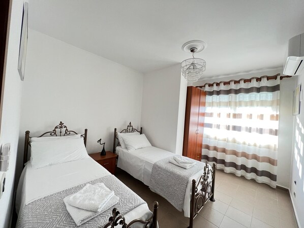 A bedroom with two single beds, a wooden wardrobe, a chandelier, and a window with striped curtains.