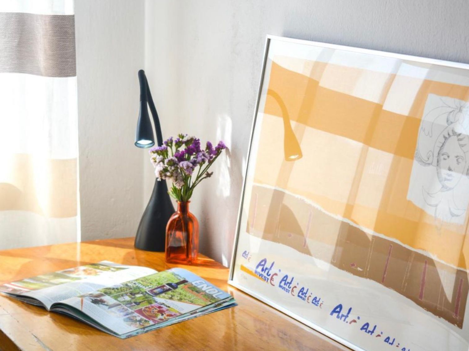 Una revista sobre una mesa de madera, un florero con flores y una obra de arte enmarcada con texto.