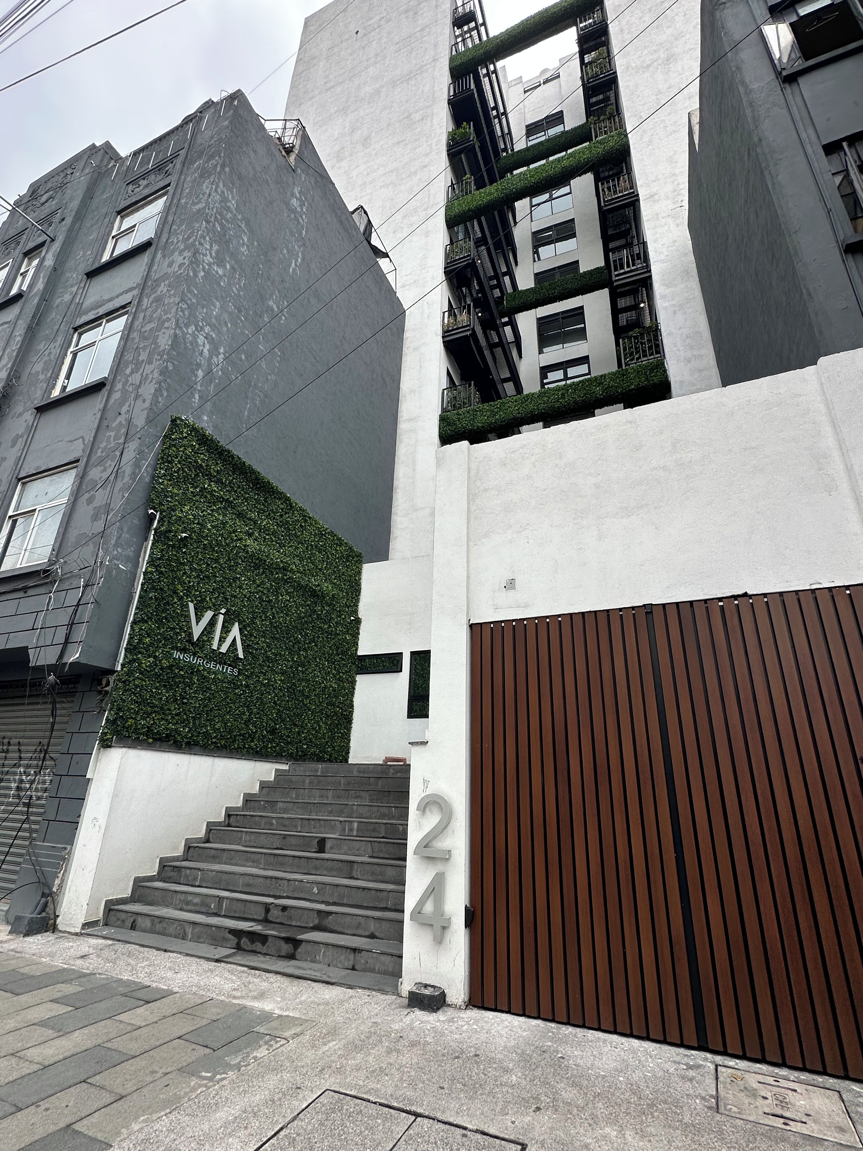 Ein modernes Gebäude mit einer großen Beschilderung mit der Nummer 24, einer Treppe, die zum Eingang führt, und einer grünen Wand mit dem Schriftzug 'VIA INSURGENTES'.