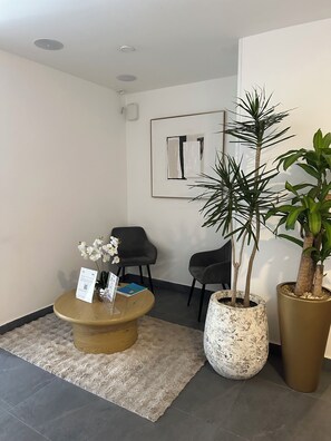 A reception area with a wooden coffee table, two chairs, a large potted plant, and a framed abstract artwork on the wall.