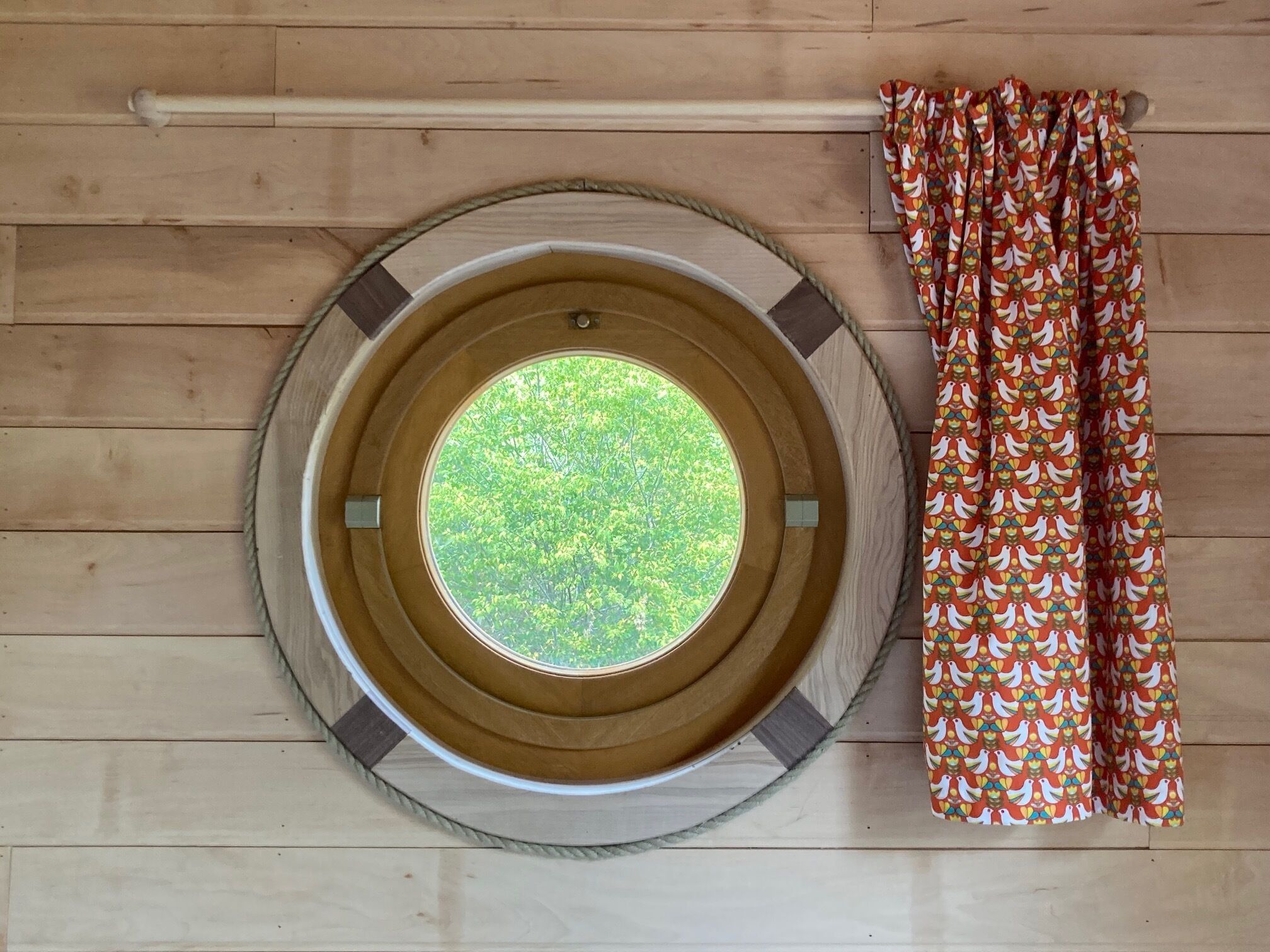 A circular window with a view of greenery, a wooden ceiling, and a curtain with a bird pattern.