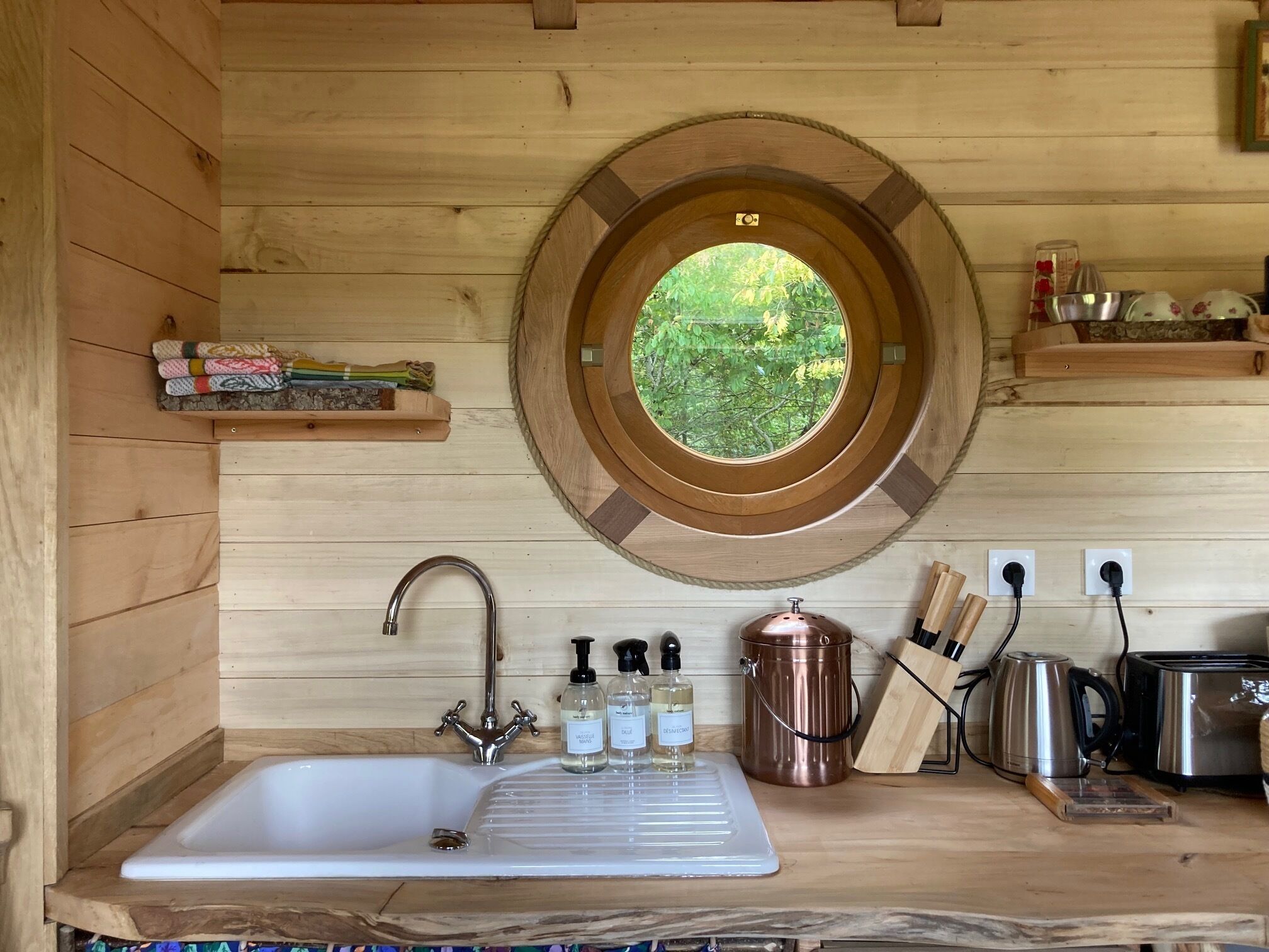 A kitchen with a wooden interior, a round window, a sink, and various kitchen utensils.