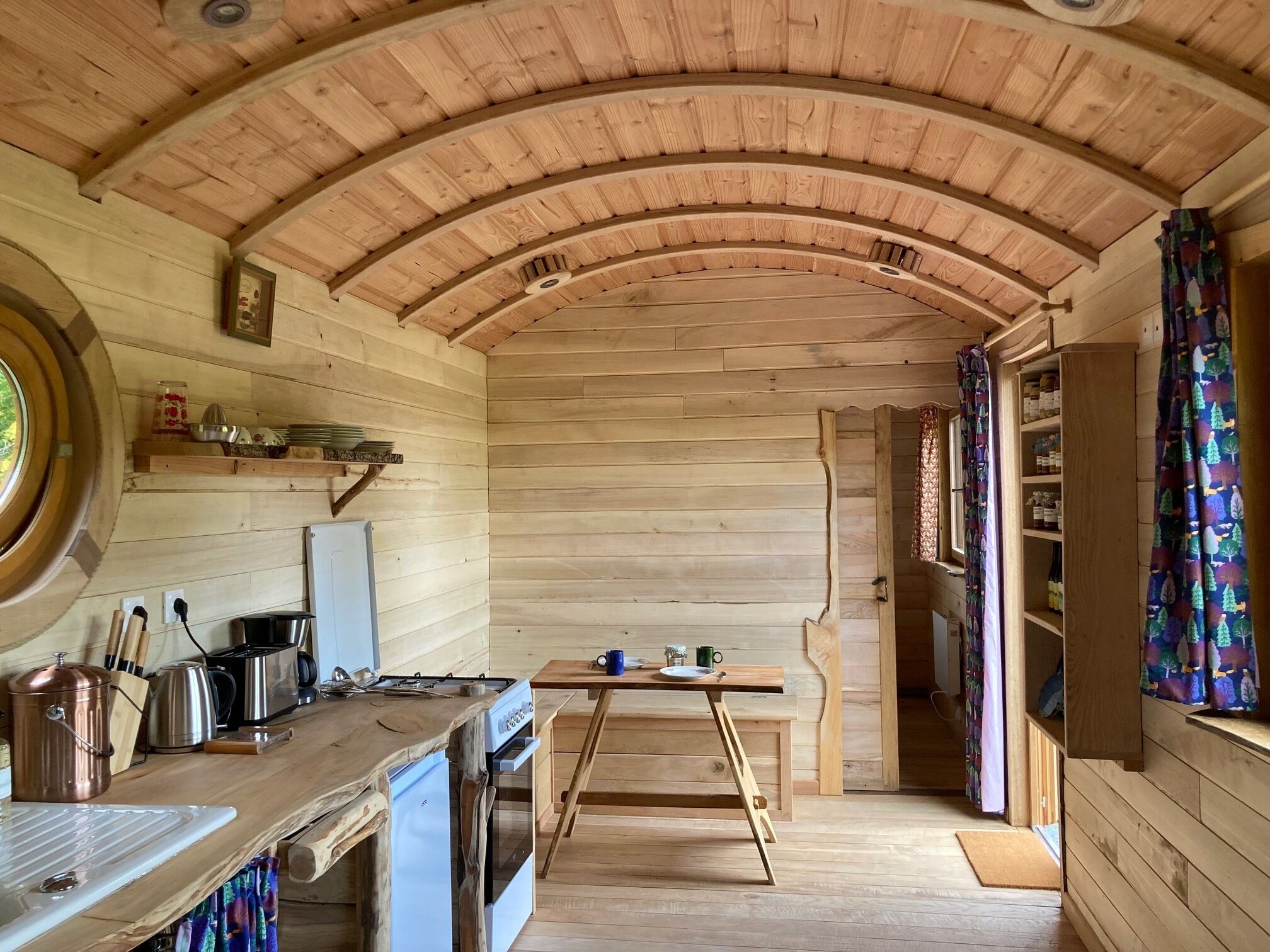 A wooden interior with a curved ceiling, a kitchen area with appliances, a dining table, and a window with curtains.