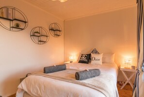 A neatly made bed with a white comforter, two pillows, and two rolled towels. There are three wall-mounted shelves with decorative items.