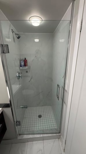A glass-enclosed shower stall with a tiled floor, a handheld showerhead, and a shelf with toiletries. - Fort Harbour Suites - Downtown Toronto (Toronto)