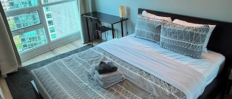 Un dormitorio moderno con una cama grande, un escritorio, una silla y vistas a edificios a través del ventanal, además de una lámpara fijada a la pared.