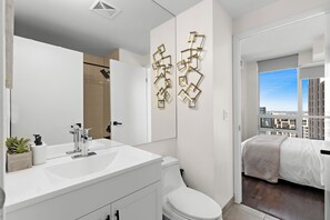 Un baño moderno con un tocador blanco, inodoro y ducha. Hay un espejo grande y arte decorativo en la pared. El baño da a un dormitorio con cama y vistas a edificios.